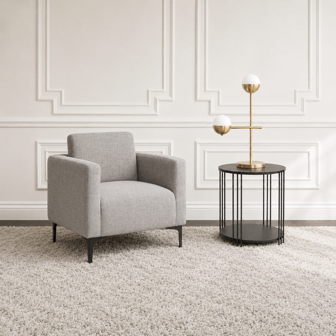 A modern light gray upholstered armchair sits on a shag rug beside a black wire side table with a gold and white two-sphere table lamp, set against a white paneled wall.