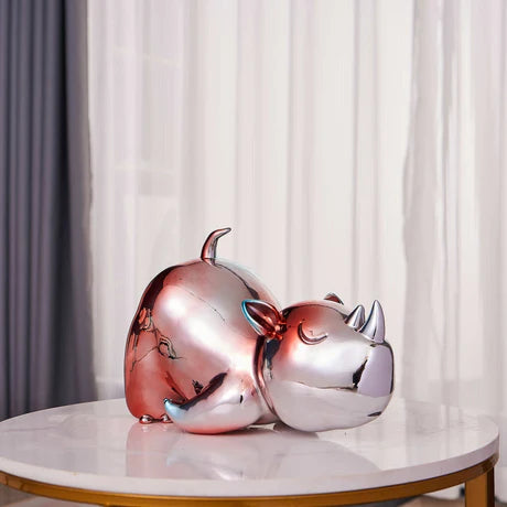 Two metallic rhino figurines on a marble surface with white curtains in the background