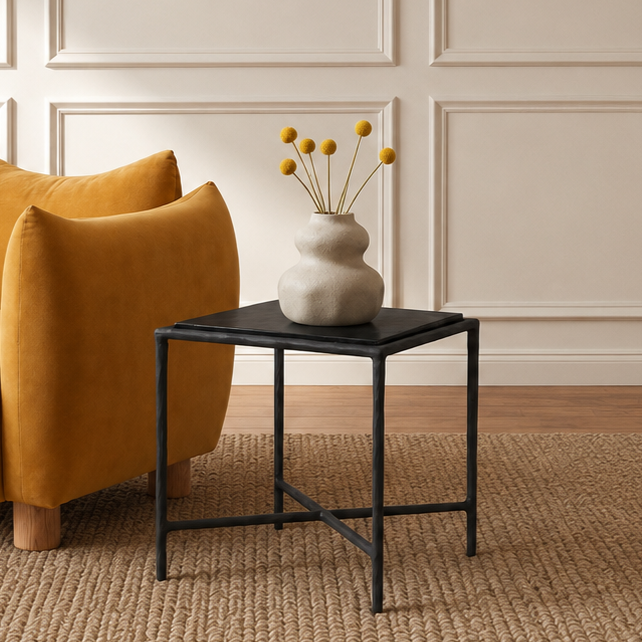 A black square metal side table with a woven top, styled in a living room next to a mustard yellow armchair. A white ceramic vase with yellow pom-pom stems sits on the table.