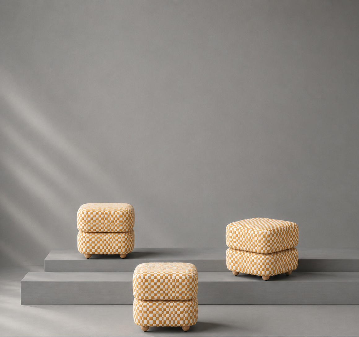 Three small woven ottomans in a light beige and brown pattern are arranged on gray pedestal steps against a gray wall with soft diagonal light rays.
