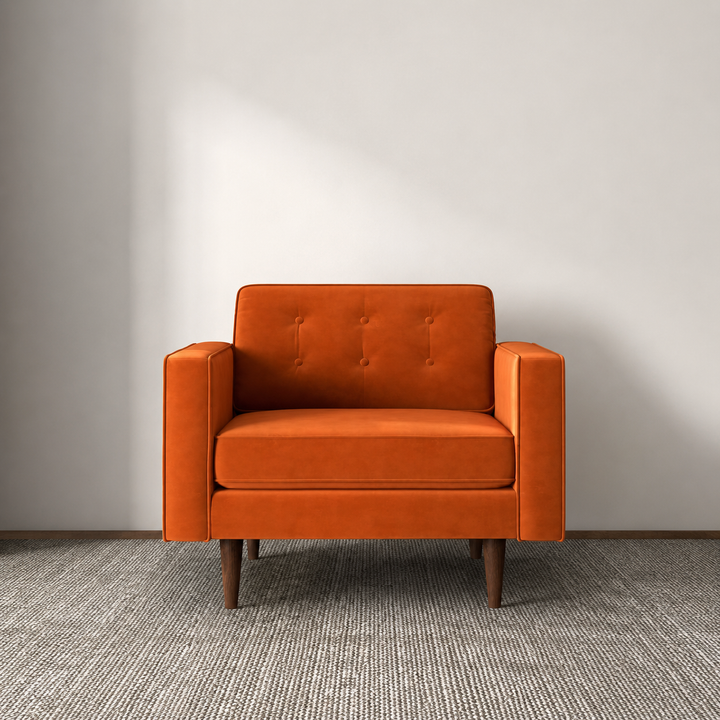An orange upholstered armchair with a tufted backrest and tapered wooden legs, centered against a plain light gray wall on a textured gray carpet.