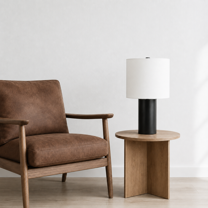 A modern table lamp with a white drum shade and black cylindrical base sits on a round wooden side table next to a brown upholstered wooden armchair against a white wall.