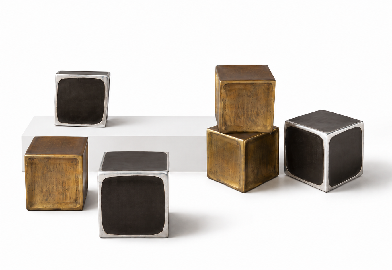 Five metallic cube-shaped decorative objects with dark rectangular faces and silver edges, arranged on and beside a white rectangular pedestal against a light gray background.