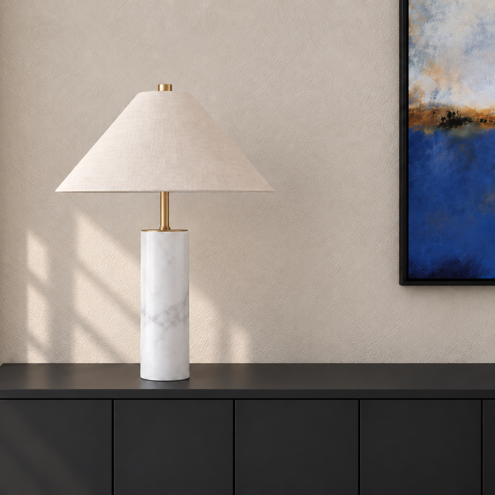 A table lamp with a white marble cylindrical base and a beige fabric conical shade, styled on a dark cabinet against a textured beige wall. 