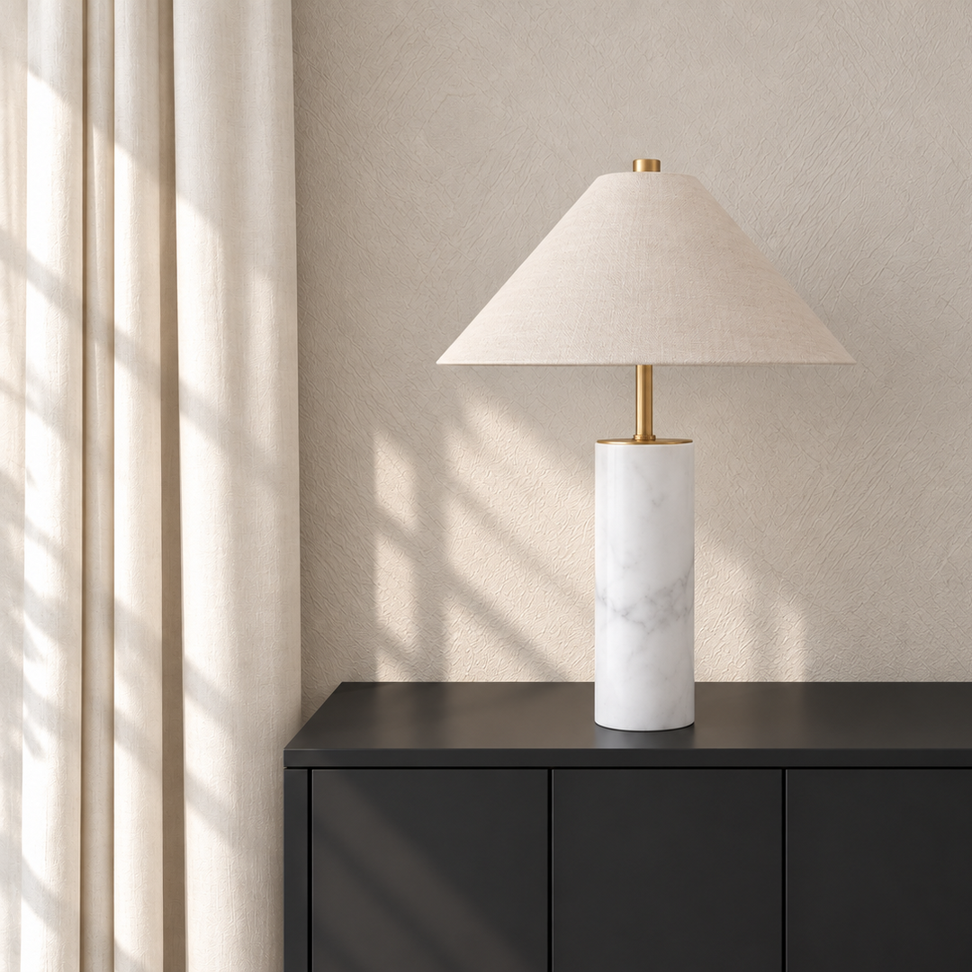 A modern table lamp with a white marble cylindrical base and a beige conical fabric shade, styled on a dark console table against a textured beige wall.  