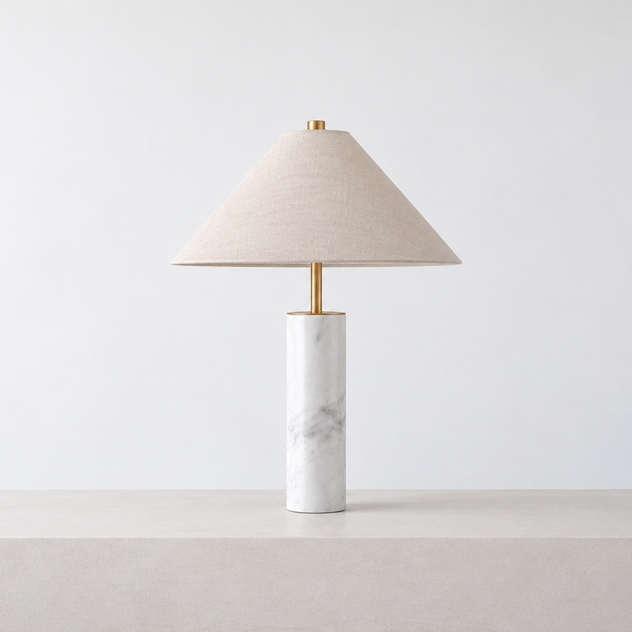 A modern table lamp with a tapered beige linen shade, a gold-finished stem, and a white marble cylindrical base, shown on a light gray pedestal against a white background.  