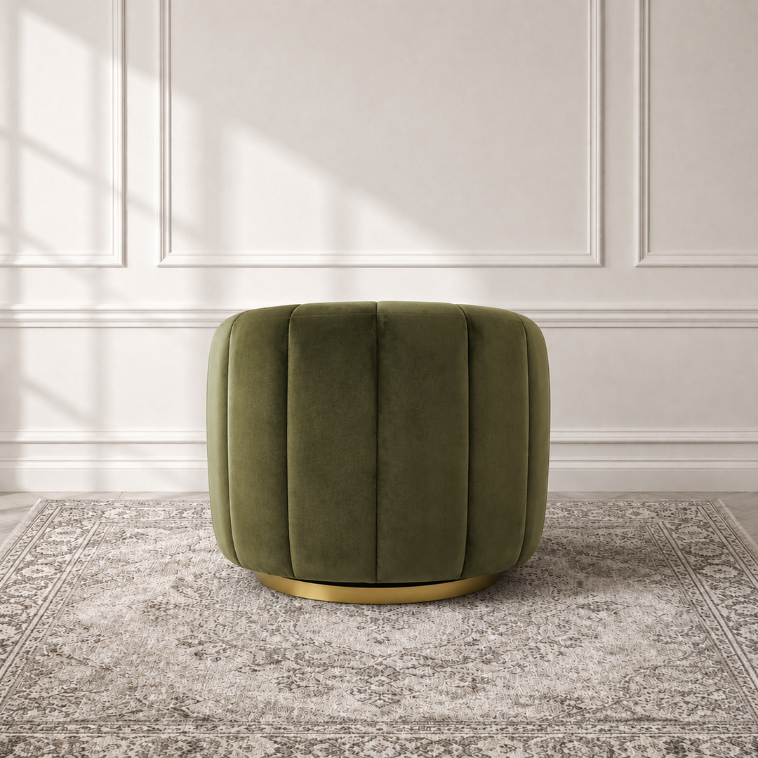 An olive green velvet ottoman with vertical channel tufting and a gold metal base, set on a gray patterned rug against a white paneled wall.