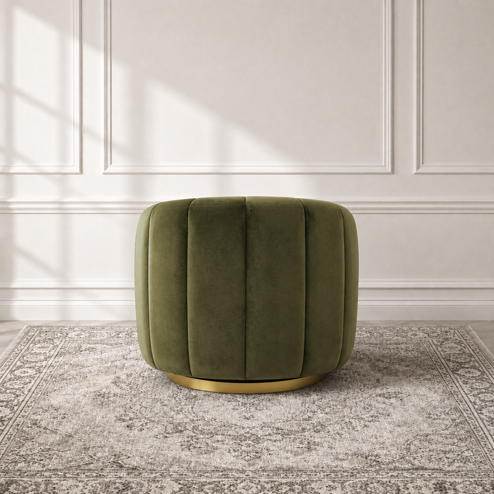 An olive green velvet ottoman with vertical channel tufting and a gold metal base, set on a gray patterned rug against a white paneled wall.