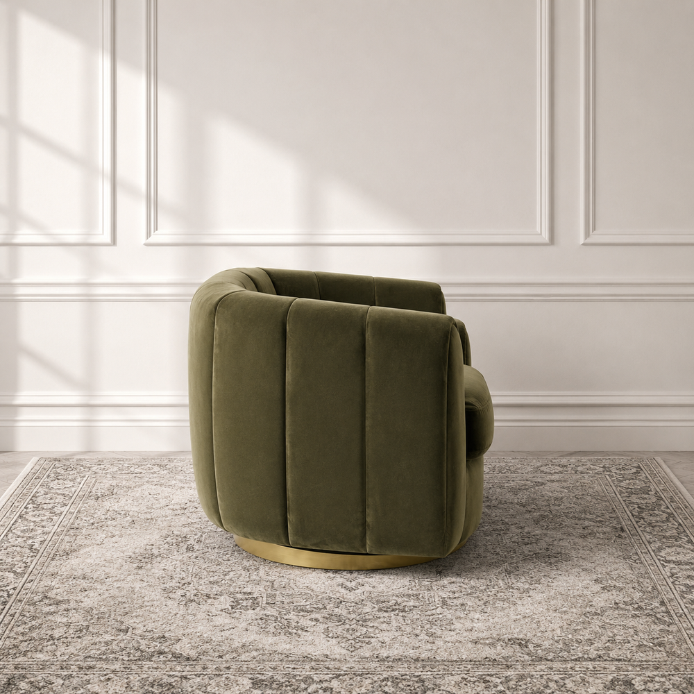 An olive green velvet swivel armchair with a gold metal base, set on a gray patterned rug against a white paneled wall with soft window shadows.