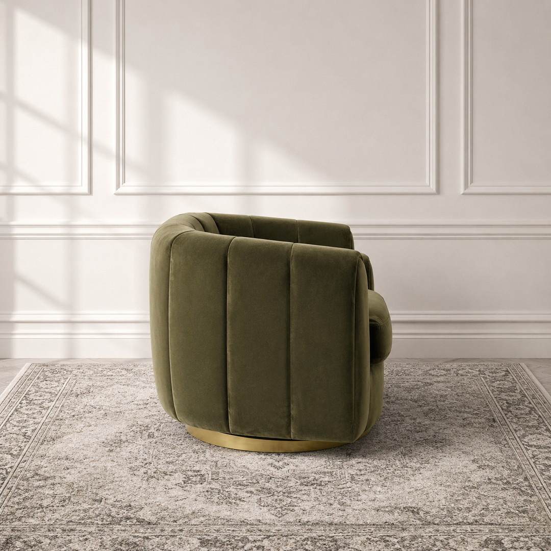 An olive green velvet swivel armchair with a gold metal base, set on a gray patterned rug against a white paneled wall with soft window shadows.