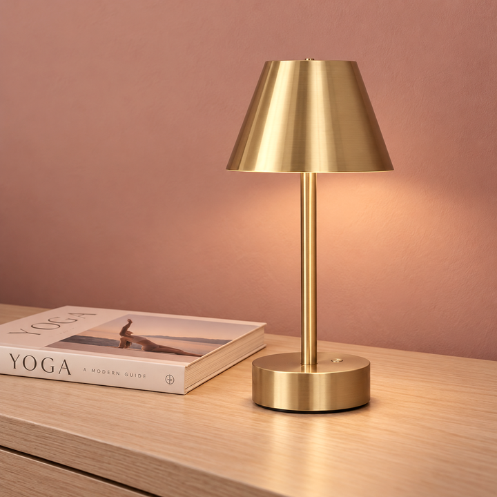 A modern table lamp with a brushed brass/gold finish, featuring a tapered shade, slim stem, and round base, shown lit on a light wood surface next to a book against a warm pinkish-brown wall. 
