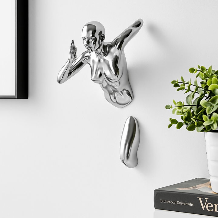 Metallic wall sculpture of a nude figure on a white wall with a plant and book in the background.