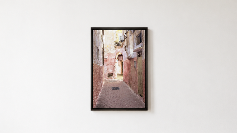 A framed vertical art print depicting a narrow old European alleyway with warm-toned buildings, hung on a white wall above a light wood floor.  