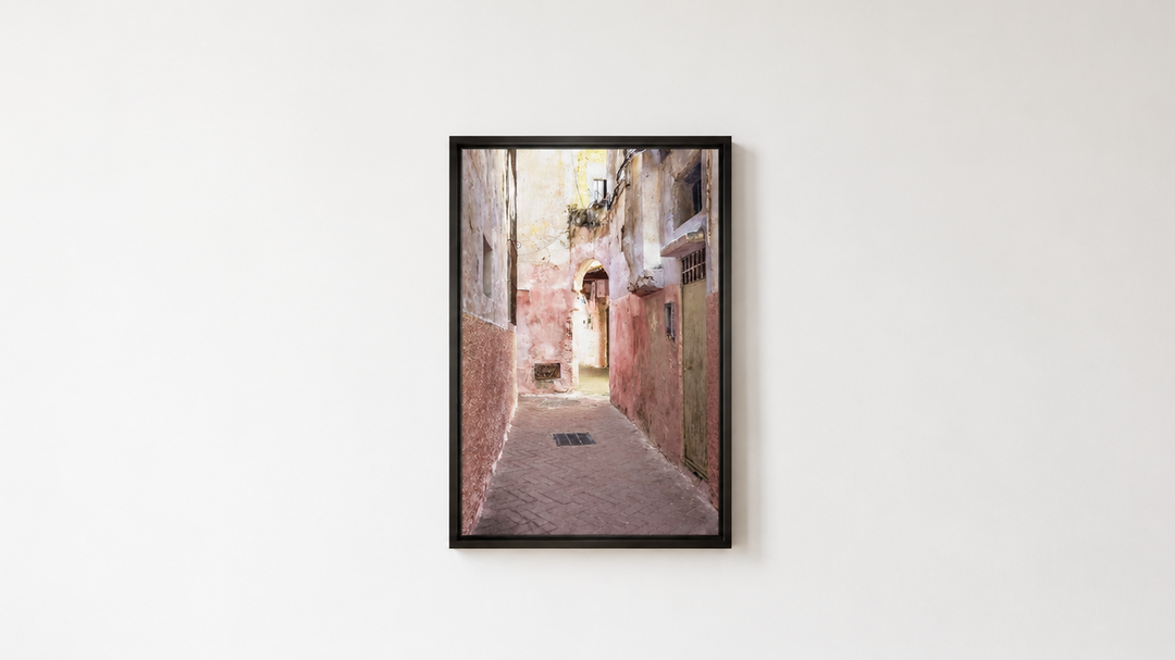 A framed vertical art print depicting a narrow old European alleyway with warm-toned buildings, hung on a white wall above a light wood floor.  