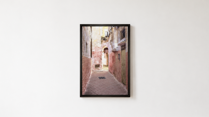 A framed vertical art print depicting a narrow old European alleyway with warm-toned buildings, hung on a white wall above a light wood floor.  