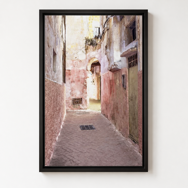 A framed vertical art print depicting a narrow old European alleyway with weathered pink and beige walls, hanging shutters, and a paved path, shown hanging on a white wall. 