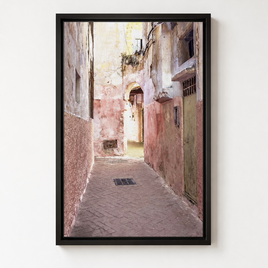 A framed vertical art print depicting a narrow old European alleyway with weathered pink and beige walls, hanging shutters, and a paved path, shown hanging on a white wall. 