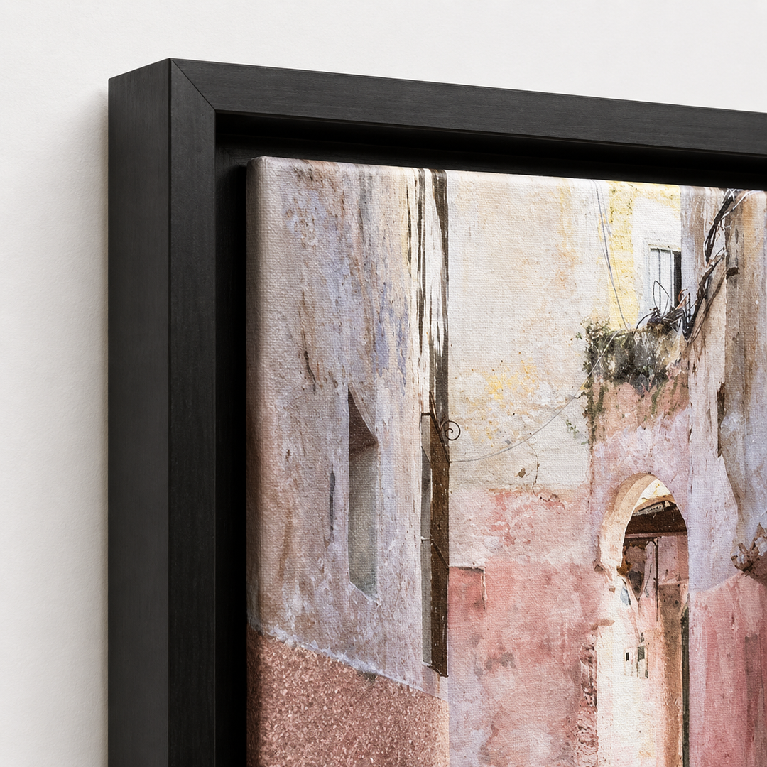 A framed wall art print showing a narrow old stone alley with weathered beige, pink, and gray buildings, presented in a black frame against a light neutral wall.