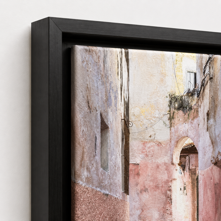 A framed wall art print showing a narrow old stone alley with weathered beige, pink, and gray buildings, presented in a black frame against a light neutral wall.