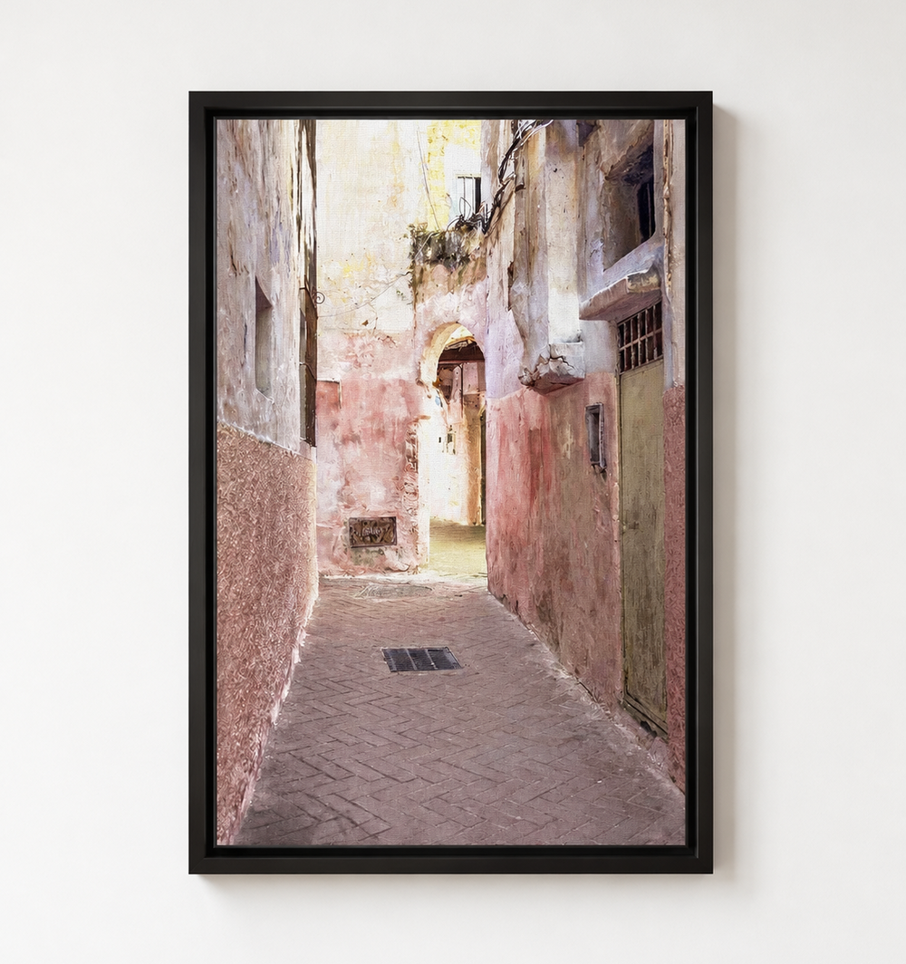 A framed vertical art print depicting a narrow old European alleyway with weathered pink and beige walls, hanging shutters, and a paved path, shown hanging on a white wall. 