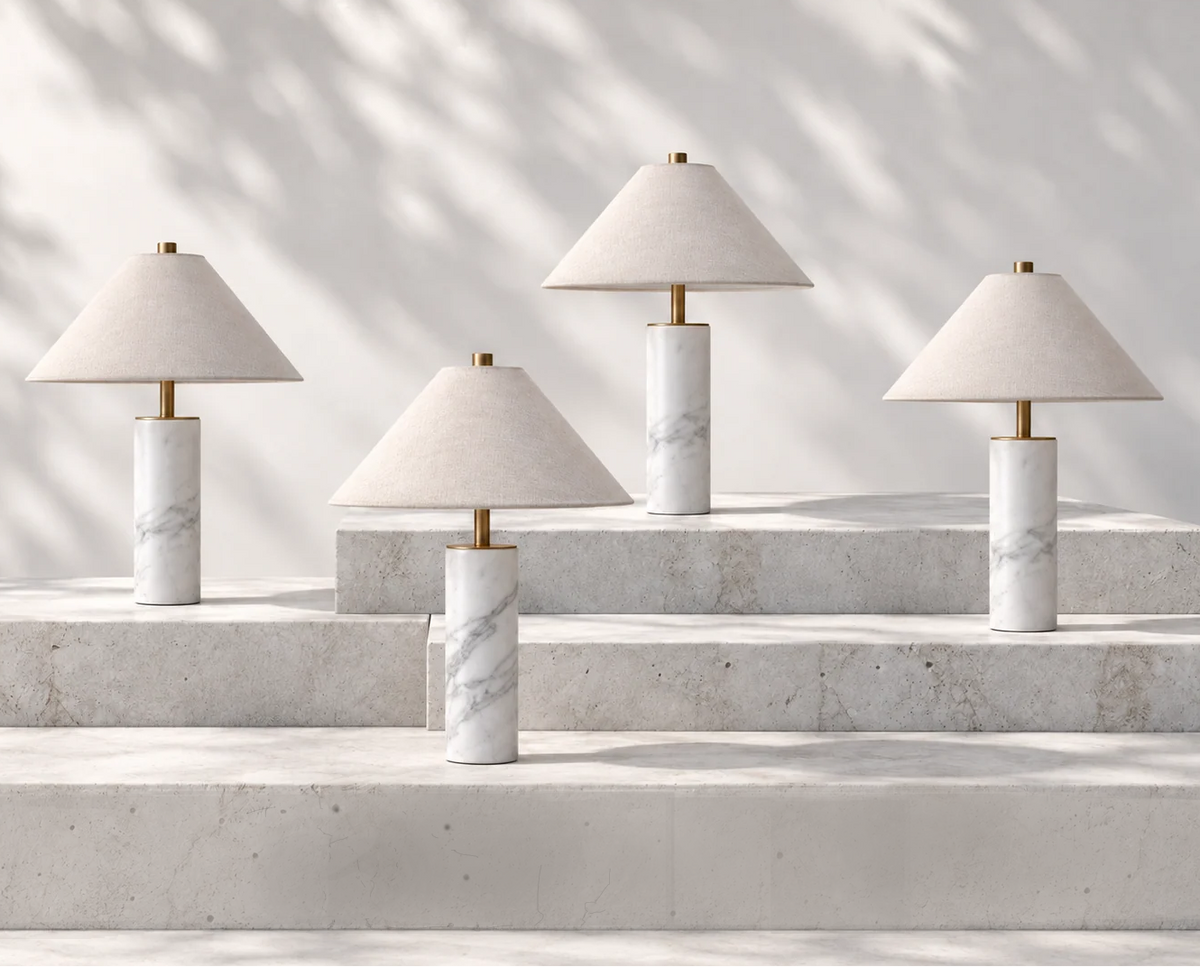 Four table lamps with tapered off-white fabric shades and cylindrical white marble bases with subtle gray veining, arranged on light stone steps against a softly shadowed wall. 