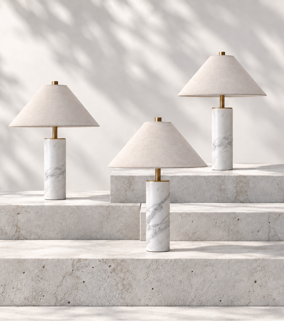 Four modern table lamps with white marble cylindrical bases and tapered off-white fabric shades, arranged on light stone steps against a softly lit wall with leaf shadows.