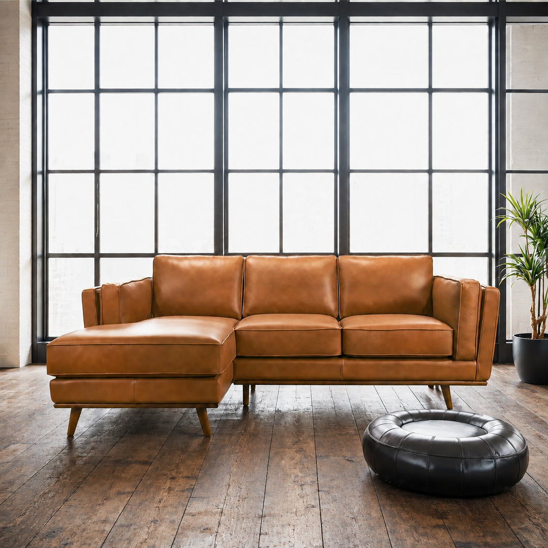 A modern sectional sofa in tan leather with a chaise lounge, positioned on a dark wood floor in front of large black-framed industrial windows. A round black leather ottoman sits in the foreground, and a potted plant is visible on the right.