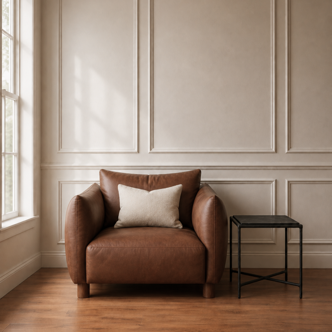 A brown leather armchair with a cream throw pillow sits in a room with beige paneled walls and a wooden floor, next to a small black side table.