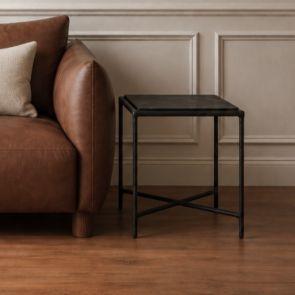 A brown leather armchair with a cream throw pillow sits in a room with beige paneled walls and a wooden floor, next to a small black side table. 