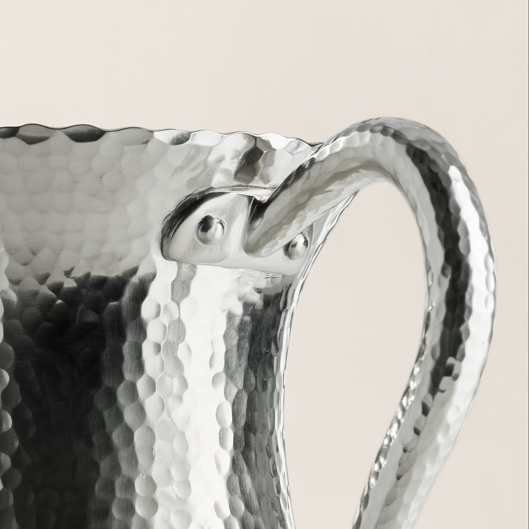 Close-up of a silver pitcher with a textured surface on a beige An artful hand-hammered technique creates the distinctive pebbled texture that defines our Evans pitcher. Made with brass, it features a lustrous nickel finish that highlights natural variations throughout. background